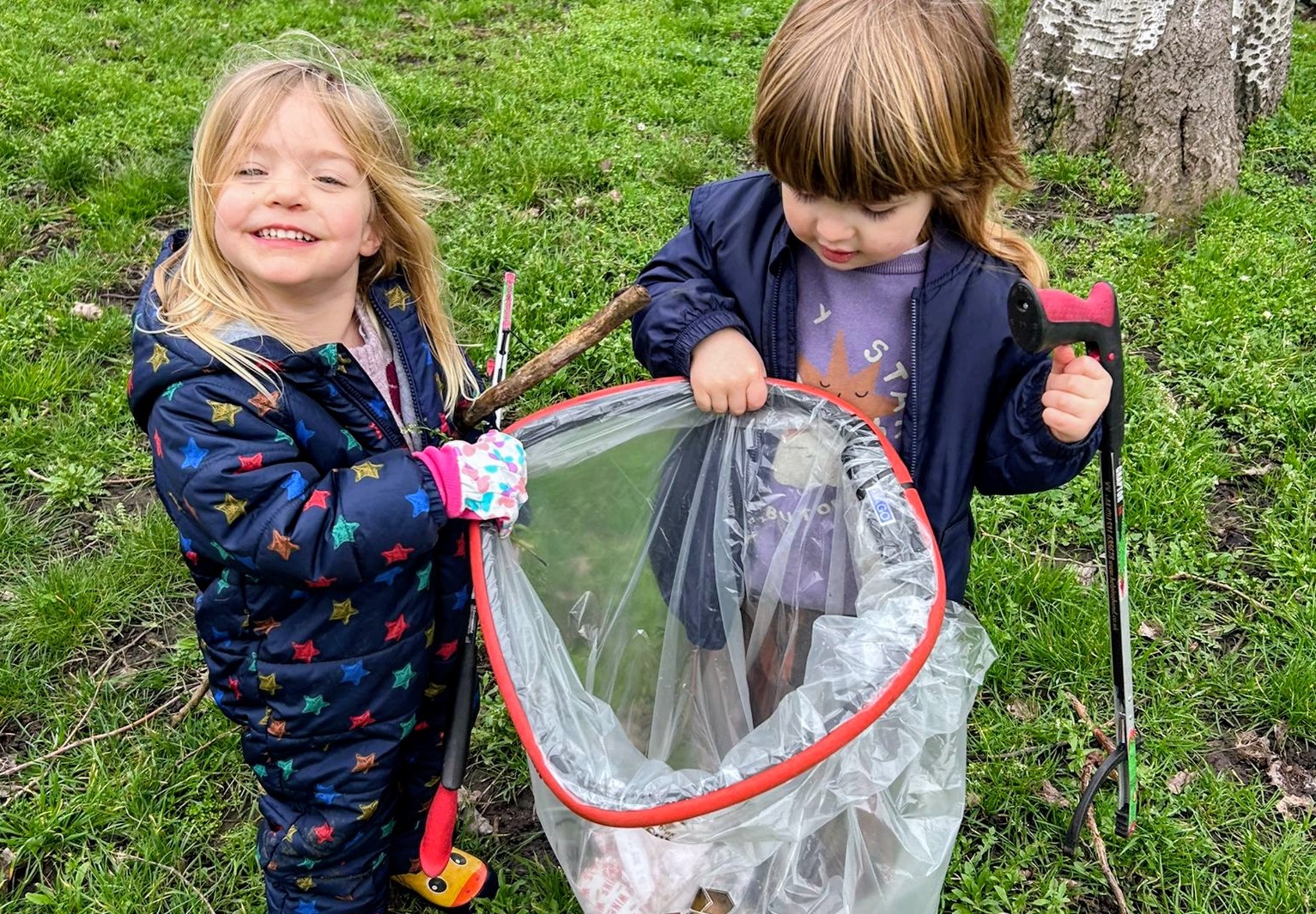 Two children holding a clear bin bag while gardening and cleaning litter, against a grassy background.