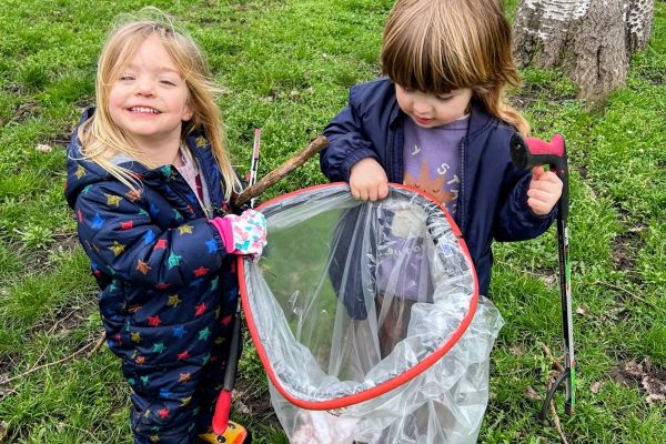 Two children holding a clear bin bag while gardening and cleaning litter, against a grassy background.